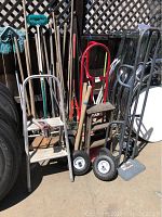 Photo showing hand trucks with metal frames featuring wooden platforms and pneumatic tires alongside yard tools like rakes, picks, and shovels with wooden handles