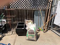 Photo showing two black metal plant stands, two black square planters, four bags of ACE Western Bark mulch, and garden tools like brooms and an auger, arranged outdoors.