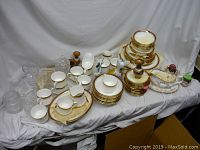 Wide view of entire lot showing stacked plates, cups, glassware, and porcelain items on white cloth.