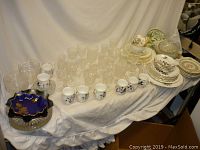 Full lot spread on white backdrop showing stacks of different china plates, glassware including clear pressed drinking glasses, vintage china cups, and decorative glass bowls.