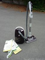 Full canister vacuum cleaner with hose and upright nozzle next to several vacuum cleaner bags still sealed in plastic packaging.
