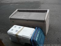 Closed view of large brown outdoor storage box, white wheeled cooler, and three blue tote lids stacked beside them on pavement.