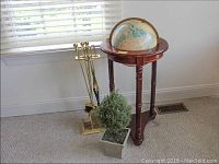 Photo showing brass fireplace tools, wooden globe stand with globe, and faux floral arrangement against a white wall and window