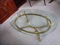 Oval metal base coffee table with clear glass top placed on carpeted floor, showing entire table and details of metal base.