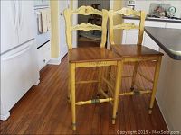 Pair of yellow distressed bar chairs with cute carved backs shown in kitchen setting on wood floor.