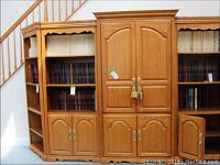 Front view of the wooden media armoire cabinet flanked by two side shelving units with decorative scalloped valances, filled with books. Shows two arched double doors with brass handles and tassels.