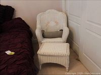 White wicker oversized armchair with cushion and matching ottoman shown beside a bed.
