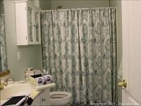View of bathroom showing fabric shower curtain with green botanical pattern, white cabinetry, and countertop with some boxed items and electronic device.