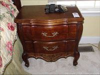Front view of the wooden bombe style nightstand showing two drawers with brass handles and carved bottom trim