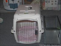 Photo of white plastic pet travel crate with metal wire door and pink cushion inside, next to clear glass fish tank with black frame, shown on carpeted floor.