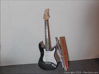 Full view of the black FirstAct electric guitar standing next to a folded metal music stand in a cardboard box against a wall.