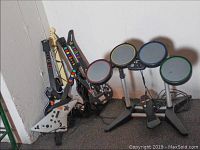 Photo showing 3 Xbox 360 guitar controllers and Xbox drum set arranged against a wall.