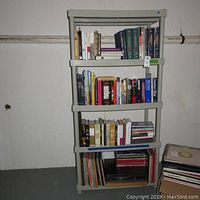 Front view of grey Keter shelving unit with four shelves holding books and records. Shows usage wear like scratches.