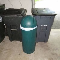 Three garbage cans shown together in a garage setting: small round green swing-top trash can in front, large black Rubbermaid and gray Toter Inc rolling bins behind.