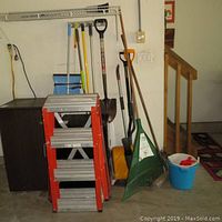 Photo of the orange 6 ft metal folding ladder with wide steps next to blue buckets, brooms, shovels, and rakes.