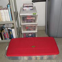 3 drawer rolling storage cart filled with VHS tapes. Large red lid plastic storage bin in front containing more tapes and DVDs.