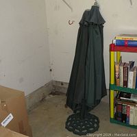 Two closed green patio umbrellas standing side by side in a garage corner next to a bookshelf and cardboard box.