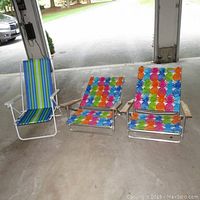 Three beach chairs. Two with multicolor pineapple pattern fabric and metal frames, folded. One blue-green-yellow striped high-back chair with wooden armrests and adjustable recline.