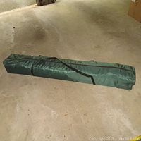 Carry case for the folding gazebo placed on a concrete floor, hunter green in color with a strap.