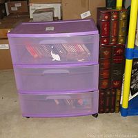 Front view of purple plastic rolling 3-drawer storage cart partially filled with CDs and wooden faux book CD holder featuring multiple faux book spines.