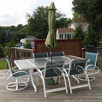 Full view of glass and metal table with 2 swivel captain chairs, 4 side chairs around it, umbrella closed in center of table on wooden deck outdoors.