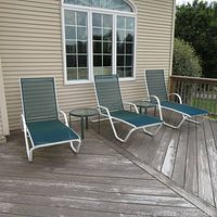 Three green mesh fabric lounge chairs with white metal frames arranged on a wooden deck in front of a window. Two round side tables with glass tops next to chairs visible. Used outdoor patio furniture.