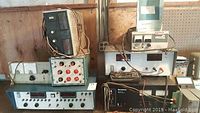 Photo of multiple power supply units stacked and arranged against wooden wall, showing front panels with meters, dials, and wires.