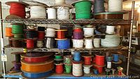 Three metal shelves holding multiple spools of wire in different sizes and colors including red, white, green, black, orange, purple, blue, and brown wires.