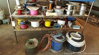 Two-tier metal shelf with multiple spools of wire in various colors and gauges; some wires on floor near spools.