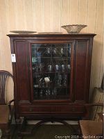 Full frontal view of the dark wood hutch showing the glass door with decorative muntins, side panels, and top molding. The hutch is filled with glassware not included.