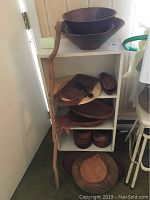 Full view of 3-tier white bookshelf holding wooden items including bowls, platters, cutting boards, small wooden spoon, wooden spool, and natural wood walking stick leaning alongside.