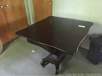 Square mahogany finished game table showing the top surface and base with casters.