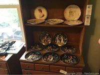Photo showing a wooden shelf with thirteen porcelain plates; the top shelf holds four assorted decorative plates with floral and classical designs, while the bottom shelves display Egyptian decorative plates mostly with black backgrounds and colorful human figure scenes.