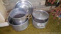 Three hand hammered aluminum stock pots grouped on floor, two with brass handles and one with aluminum handles and a lid.