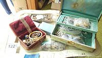 Photo showing collection of costume jewelry in red velvet box and green oriental-style jewelry box atop tray tables.