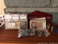 Photo of foreign coins stored in a plastic organizer with compartments, accompanied by two plastic bags with loose coins and two paper rolls of coins, all placed on a wooden surface in front of a stone wall.
