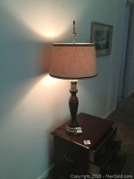 Lamp illuminated on wooden furniture against wall, showing full lamp with beige fabric shade.