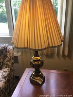 Brass table lamp lit showing beige pleated shade, on wooden side table near window with floral couch nearby.