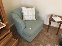 Small light green rocker swivel chair with a white patterned pillow on it, placed on carpeted floor next to a small wooden table.
