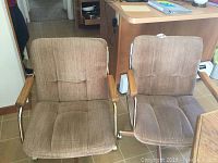 Two light brown fabric swivel chairs with oak armrests and wheels shown on tiled floor near wooden furniture.
