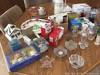 Wide view of assorted items on wooden dining table including candlesticks, candles, decorative platters, tea kettle box, thermos, and lightbulbs.
