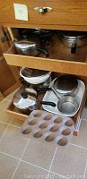 Drawer with multiple stainless steel pots and pans with black handles, metal lids, muffin pan in foreground