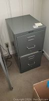 Full view of the gray 2-drawer metal filing cabinet against wall on carpet