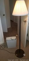 Floor lamp and paper shredder shown on carpeted floor near door, lamp turned on showing pleated shade and slim, dark pole with circular base.
