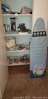 A closet shelving unit displaying various household items including an ironing board with a decorative cover featuring flowers and tulips, a small steam iron box, and sewing supplies.
