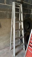 6ft aluminum step ladder with four wide steps leaning against a cinder block wall indoors.