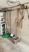 Wide view showing garden tools hanging on a wall including shovels, pitchfork, garden rake, a wooden basket, soil bag, and coiled green garden hoses on floor.