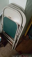 Four stacked Samsonite metal folding chairs with beige frames and green vinyl padded seats and backs, showing wear and weathering.