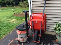 Wide view of the Rigid wet dry vac and the red Craftsman wheelbarrow positioned next to a house exterior wall.