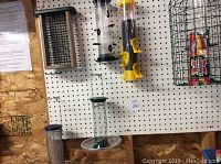 Six bird feeders displayed on wall hooks against pegboard panel, showing variety of styles and sizes.